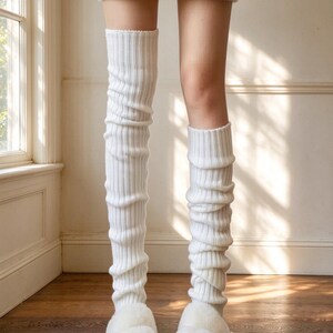 Op de afbeelding: Een paar witte geribbelde gebreide beenwarmers die tot boven de knieën reiken, gecombineerd met witte pluche kunstbont slippers. De beenwarmers hebben een verticale ribbelstructuur en een zachte pasvorm, terwijl de slippers een dik, pluizig bandontwerp hebben.