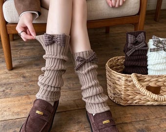 Könnte beinhalten: Ein Paar beigefarbene, gerippte Strickstulpen mit einer braun-weißen karierten Schleife am oberen Rand. Die Stulpen werden über den Unterschenkeln getragen und mit dunkelbraunen Wildleder-Loafern kombiniert, die vorne einen goldenen Metallakzent haben.