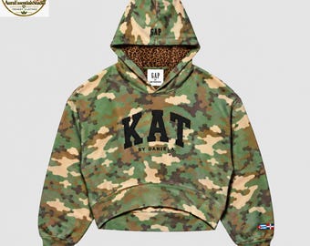 May include: A cropped camouflage hoodie featuring a green, brown, and tan pattern. The front displays the black text KAT BY DANIELA. The hood interior has a brown leopard print, and the sleeve features small Cuban and Dominican flag icons.