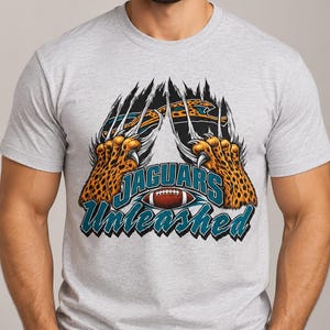 May include: A light gray short-sleeve t-shirt featuring a graphic of two leopard paws with sharp claws tearing through the fabric. The design includes the text "JAGUARS Unleashed" in teal and black lettering with a brown football in the center.