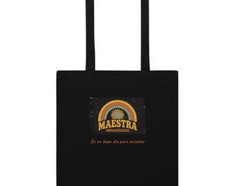 May include: A black tote bag featuring a graphic with a sun and rainbow arch above the text MAESTRA ORGANIZADA. Below the graphic, the text reads Es un buen día para enseñar. The design has a distressed, weathered texture.