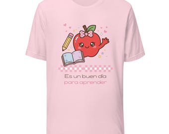 May include: A light pink t-shirt featuring a graphic of a smiling red apple with a bow, a pencil, and an open book. Below the graphic is a pink and white checkered pattern and the text Es un buen día para aprender in dark lettering.