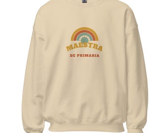 May include: A cream-colored crewneck sweatshirt featuring a graphic of a multi-colored rainbow and a smiling sun. Below the graphic, the text reads MAESTRA in large yellow letters and DE PRIMARIA in smaller red letters.
