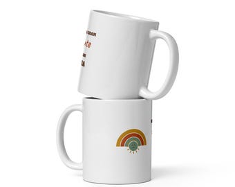 May include: Two stacked white ceramic mugs. The top mug features brown and red text that reads A GRAN mente UN GRAN DIA. The bottom mug displays a small graphic of a multi-colored rainbow in shades of orange, yellow, and green with a smiling sun.