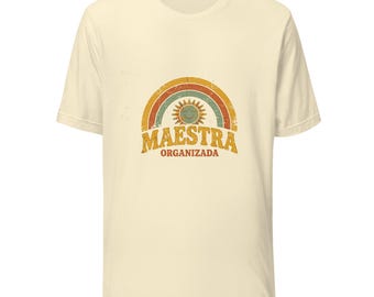 May include: A cream-colored short-sleeve t-shirt featuring a distressed graphic of a rainbow and a sun. Below the graphic, the text reads "MAESTRA ORGANIZADA" in a bold, weathered font. The design uses muted tones of orange, teal, and yellow.