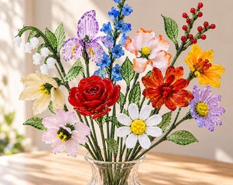 May include: A bouquet of handcrafted glass flowers in a clear glass vase. The arrangement features a red rose, white daisy, purple iris, lily of the valley, blue bell-shaped flowers, and red berries, all with green glass leaves and stems.