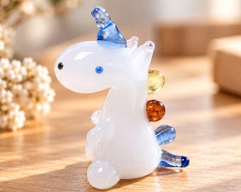 May include: A small glass figurine of a white creature with a blue horn, a black eye, and a blue eye. The creature features a yellow sphere, an amber sphere, and blue accents along its back and tail, standing in a upright position.