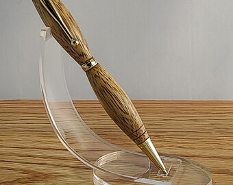 May include: A wooden pen with gold accents is displayed on a clear acrylic pen stand. The pen has a brown wood grain pattern and a gold-colored clip and tip.