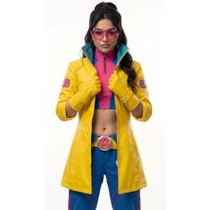 May include: A bright yellow long coat with teal lapels and matching yellow gloves, worn over a pink and blue cropped top and blue trousers. The outfit includes a pink belt with a prominent letter A buckle and pink-tinted visor sunglasses.