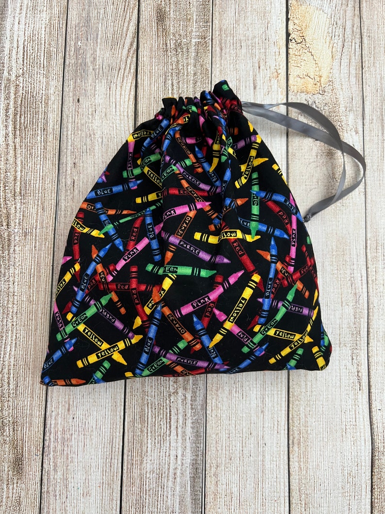 Pode incluir: Uma bolsa de cord&atilde;o preto com um padr&atilde;o de giz de cera colorido. Os giz de cera est&atilde;o dispostos em diagonal e t&ecirc;m as palavras "vermelho", "azul", "amarelo", "verde", "roxo", "laranja" e "rosa" escritas neles.