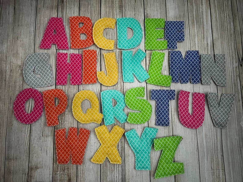 Pode incluir: Um conjunto de letras do alfabeto de feltro em v&aacute;rias cores com um padr&atilde;o de diamante. As letras est&atilde;o dispostas em ordem alfab&eacute;tica sobre um fundo de madeira.