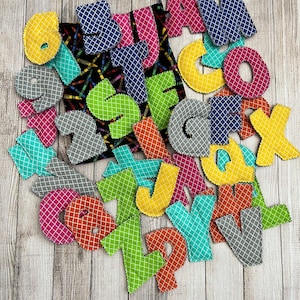 Pode incluir: Um conjunto de letras do alfabeto de tecido colorido em v&aacute;rias cores, incluindo amarelo, verde, azul, rosa, laranja e cinza. As letras est&atilde;o dispostas em uma pilha sobre uma superf&iacute;cie de madeira.