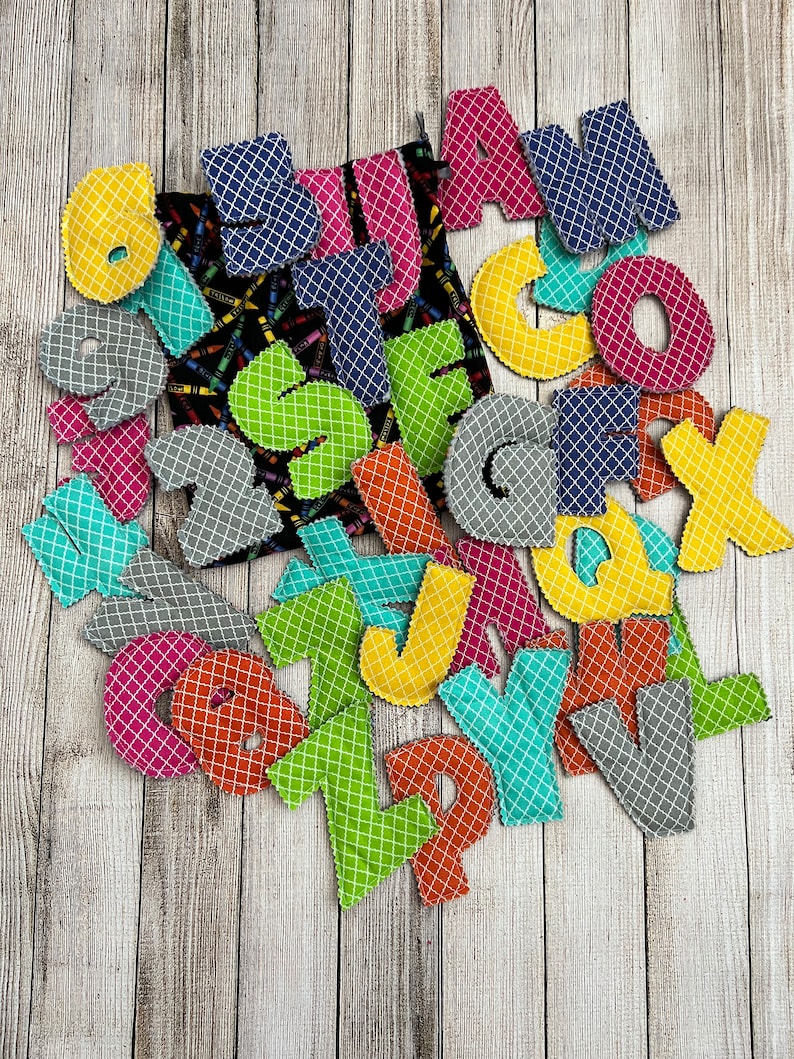 Pode incluir: Um conjunto de letras do alfabeto de tecido colorido em v&aacute;rias cores, incluindo amarelo, verde, azul, rosa, laranja e cinza. As letras est&atilde;o dispostas em uma pilha sobre uma superf&iacute;cie de madeira.