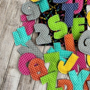 Pode incluir: Um conjunto de letras do alfabeto de feltro coloridas com um padr&atilde;o geom&eacute;trico. As letras s&atilde;o de v&aacute;rias cores, incluindo verde, azul, amarelo, laranja, rosa e cinza. As letras est&atilde;o dispostas em uma pilha sobre uma superf&iacute;cie de madeira.