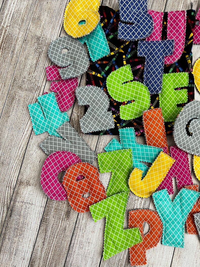 Pode incluir: Um conjunto de letras do alfabeto de feltro coloridas com um padr&atilde;o geom&eacute;trico. As letras s&atilde;o de v&aacute;rias cores, incluindo verde, azul, amarelo, laranja, rosa e cinza. As letras est&atilde;o dispostas em uma pilha sobre uma superf&iacute;cie de madeira.