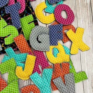 Pode incluir: Um conjunto de letras do alfabeto de feltro coloridas com um padr&atilde;o de grade branca. As letras s&atilde;o de v&aacute;rias cores, incluindo amarelo, azul, verde, rosa, laranja e cinza. As letras est&atilde;o dispostas em uma pilha sobre uma superf&iacute;cie de madeira.