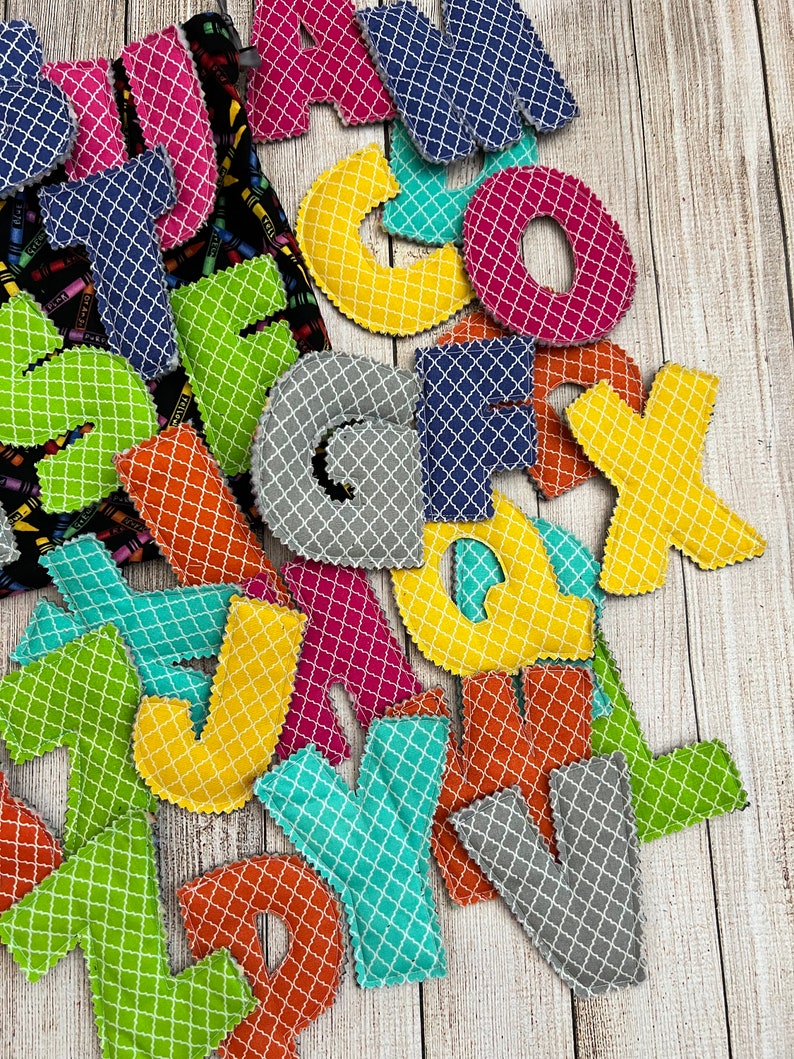 Pode incluir: Um conjunto de letras do alfabeto de feltro coloridas com um padr&atilde;o de grade branca. As letras s&atilde;o de v&aacute;rias cores, incluindo amarelo, azul, verde, rosa, laranja e cinza. As letras est&atilde;o dispostas em uma pilha sobre uma superf&iacute;cie de madeira.