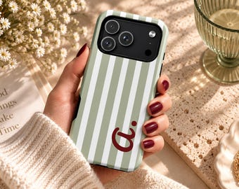Puede incluir: Una funda para smartphone con un patrón de rayas verticales en verde salvia y blanco. La funda incluye un recorte negro para el módulo de la cámara y una letra a minúscula de color rojo oscuro con un punto en la parte inferior derecha.