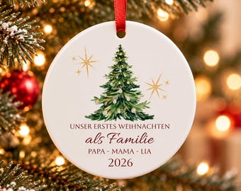 Puede incluir: Un adorno navideño redondo de cerámica blanca colgado de una cinta roja. Presenta un pino verde en acuarela, estrellas doradas y el texto: UNSER ERSTES WEIHNACHTEN als Familie PAPA - MAMA - LIA 2026 impreso en letras de color rojo oscuro.