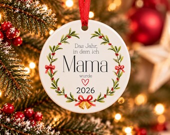 Puede incluir: Un adorno navideño redondo de cerámica blanca colgado de una cinta roja. Presenta una corona de muérdago verde con lazos rojos y campanas doradas. El texto dice: Das Jahr, in dem ich Mama wurde 2026, con un pequeño icono de corazón rojo.