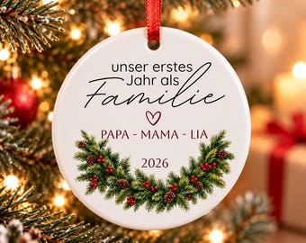 Puede incluir: Un adorno navideño redondo de cerámica blanca colgado de una cinta roja. Presenta el texto "unser erstes Jahr als Familie", "PAPA - MAMA - LIA" y "2026", junto a un icono de corazón y una corona impresa de ramas de pino verde y bayas rojas.