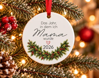 Puede incluir: Un adorno navideño redondo de cerámica blanca colgado de una cinta roja. El adorno presenta el texto en alemán "Das Jahr, in dem ich Mama wurde" sobre el año "2026" y un pequeño corazón rojo, centrado sobre una corona de pino.