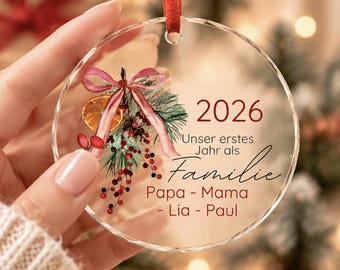 Puede incluir: Un adorno navideño redondo de acrílico transparente colgado de una cinta roja. Presenta una ilustración impresa de una rama de pino, bayas rojas y una rodaja de naranja, con el texto 2026, Unser erstes Jahr als Familie, Papa, Mama, Lia, Paul.