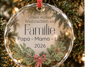 Puede incluir: Un adorno navideño redondo de cristal transparente con borde facetado, colgado de una cinta roja. Presenta el texto en alemán "Unser erstes Weihnachten als Familie Papa - Mama - Lia 2026" y una ilustración impresa de ramas de pino verdes con un lazo rojo.