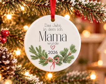 Puede incluir: Un adorno navideño redondo de cerámica blanca colgado de una cinta roja. El texto "Das Jahr, in dem ich Mama wurde" aparece con un pequeño corazón rojo y el año "2026", rodeado por una ilustración de ramas de pino y un lazo rojo.