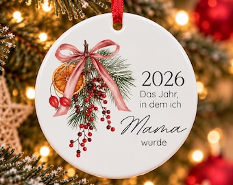 Puede incluir: Un adorno navideño redondo de cerámica blanca con cinta roja, que presenta una ilustración de ramas de pino, bayas rojas y una rodaja de naranja seca, con el texto 2026 Das Jahr, in dem ich Mama wurde.