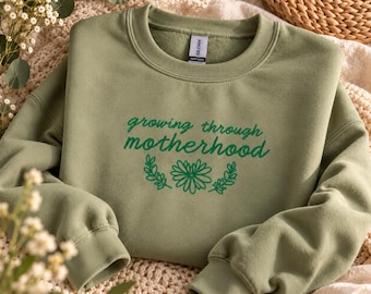 Puede incluir: Una sudadera de cuello redondo en color verde salvia con el texto bordado en verde oscuro "growing through motherhood" sobre un diseño floral de líneas simples. La prenda tiene mangas largas con puños acanalados y etiqueta Gildan.