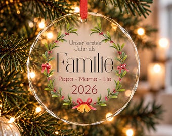 Puede incluir: Un adorno navideño redondo de cristal facetado que cuelga de una cinta roja. Presenta una corona impresa de muérdago verde con bayas rojas y un lazo. El texto dice: Unser erstes Jahr als Familie, Papa - Mama - Lia, 2026.