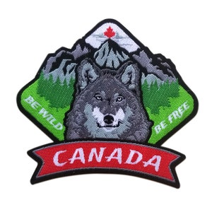 Peut inclure: Patch brodé représentant un loup gris devant une chaîne de montagnes vertes surmontée d'une feuille d'érable rouge. Le texte "Be Wild" et "Be Free" se trouve de chaque côté du loup. Une bannière rouge avec le mot "CANADA" se trouve en bas.