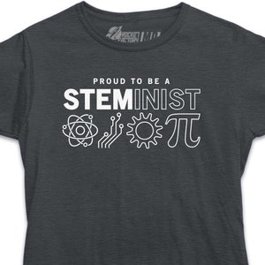 May include: A dark grey t-shirt with the text "PROUD TO BE A STEMINIST" in white. The text is accompanied by a white atom, circuit board, sun, and pi symbol.
