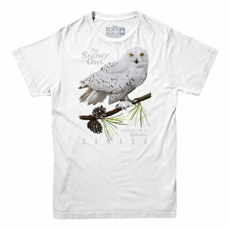 May include: White t-shirt with a snowy owl illustration. The owl is perched on a branch with pine needles and a pine cone. The text "The Snowy Owl" is above the owl and "Provincial Bird of Quebec CANADA" is below the owl.