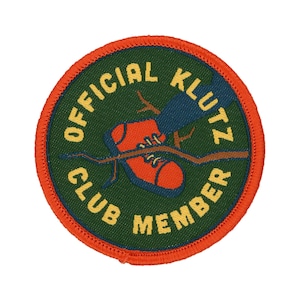May include: A round, orange-bordered patch with green fabric in the centre. The patch features a cartoon illustration of a red shoe caught on a branch, with the text "OFFICIAL KLUTZ CLUB MEMBER" in yellow lettering.