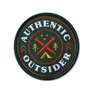 May include: Green embroidered patch with white text that reads "AUTHENTIC OUTSIDER" in a circle. The centre of the patch features a red and orange tent with crossed axes and pine trees.
