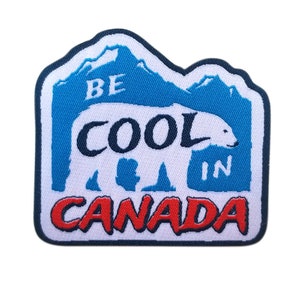Peut inclure: Un écusson rectangulaire avec une illustration d'ours polaire blanc et le texte "BE COOL IN CANADA". L'écusson a une bordure bleue avec un motif de montagne en haut et du texte rouge en bas.