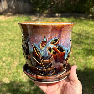 May include: A ceramic planter with a matching saucer, featuring a deep brown glaze with blue and purple drip accents. The body of the planter is decorated with intricate, cut-out leaf patterns that allow light to pass through the vessel.