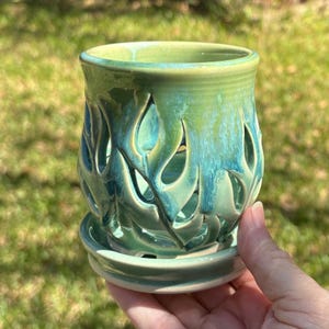 May include: A ceramic planter with a glossy glaze in shades of light green and teal. The cylindrical body features intricate, flame-shaped cutouts throughout the sides, and the piece includes a matching circular saucer base.