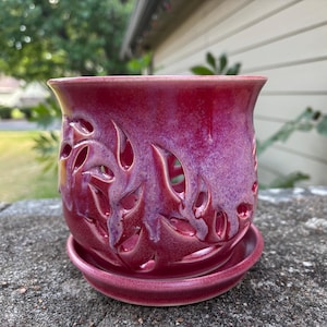 May include: A cylindrical ceramic planter with a vibrant magenta and purple glaze, featuring intricate cut-out leaf patterns throughout the body. The planter sits securely on a matching circular saucer, creating a cohesive two-piece set.