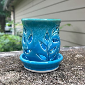 May include: A cylindrical ceramic candle holder or planter featuring a vibrant turquoise and teal gradient glaze. The exterior is decorated with intricate, cut-out leaf patterns that allow light to pass through, resting on a matching circular base.