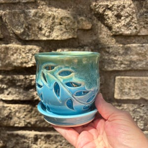 May include: A ceramic plant pot with a matching saucer, featuring a vibrant blue and green ombre glaze. The exterior of the pot is decorated with intricate, cut-out leaf patterns that reveal the interior space, creating a decorative effect.