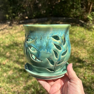May include: A ceramic planter with a glossy teal and green glaze featuring intricate cut-out leaf patterns throughout the body. The vessel includes a matching circular saucer base, showcasing a smooth finish and a rounded, bulbous shape.
