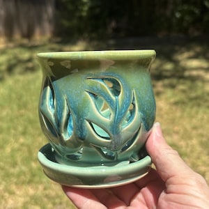 May include: A ceramic planter with a glossy finish in shades of green and blue. The vessel features intricate, leaf-shaped cutouts throughout its body, allowing for ventilation. It sits on a matching circular saucer, creating a cohesive unit.