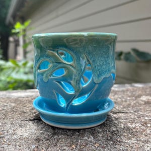 May include: A cylindrical ceramic planter with a vibrant turquoise and teal glaze. The exterior features intricate, organic cutouts throughout the body, allowing light to pass through. It sits on a matching circular saucer base with a smooth finish.