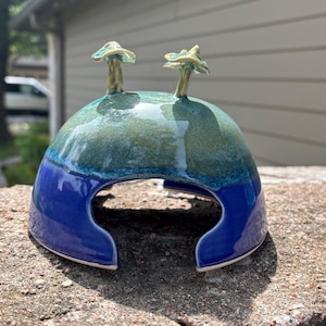 May include: A ceramic mushroom house with a blue and green glazed finish. Two small mushroom-shaped accents are on top. The house has a rounded shape with a large opening at the bottom.