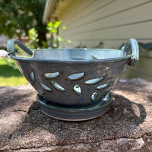 May include: A ceramic berry bowl with a glossy blue-grey glaze, featuring cut-out leaf-shaped drainage holes, two side handles, and a matching circular base plate. The bowl has a smooth, rounded rim and a deep basin for holding produce.