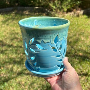 May include: A cylindrical ceramic planter with a matching saucer, featuring a vibrant turquoise glaze and a decorative cutout leaf pattern. The rim displays a lighter green drip effect, and the body has a smooth, glossy finish.