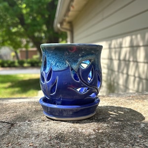 May include: A handcrafted ceramic candle holder in shades of blue. The holder features leaf-shaped cutouts and a matching saucer. The top is a lighter blue, transitioning to a deep blue at the base. A unique decorative piece.